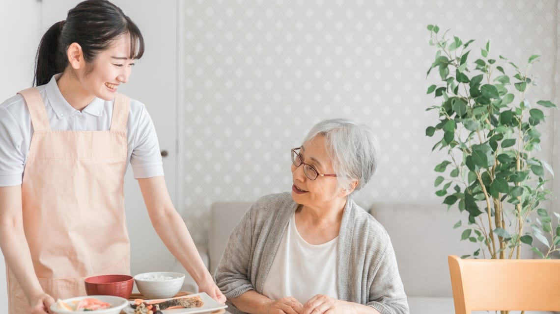 食事を運ぶ職員と、テーブルで待つ高齢の女性のイメージ
