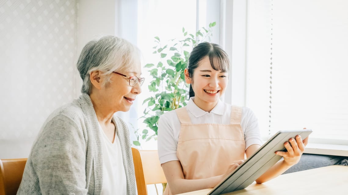 介護の専門職と、タブレットで退院後に使う介護サービスを確かめる高齢の女性のイメージ