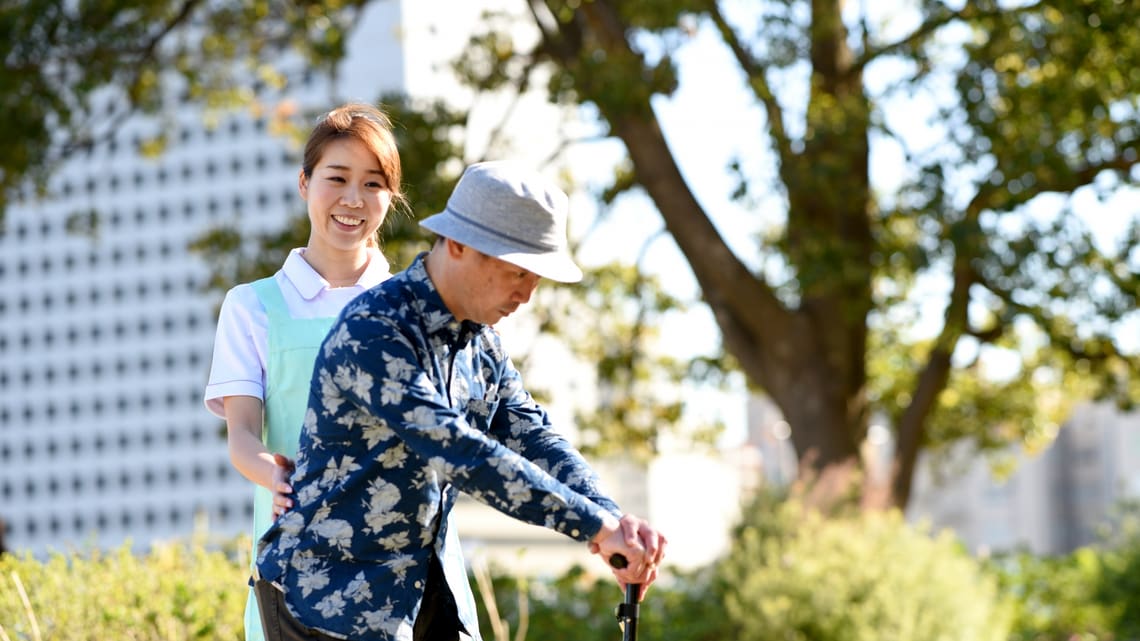 職員の付き添いで、杖をついて歩く練習をする高齢の男性のイメージ