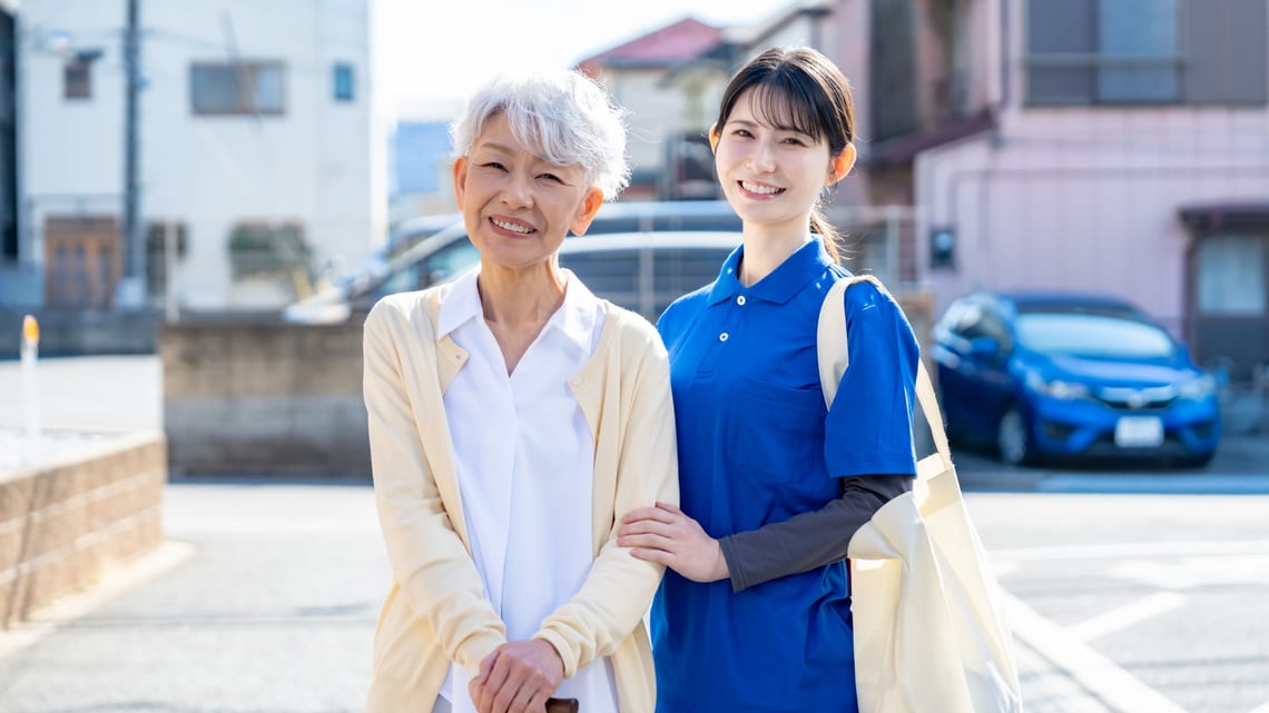 杖をついて歩く高齢の女性に、職員が付き添うイメージ