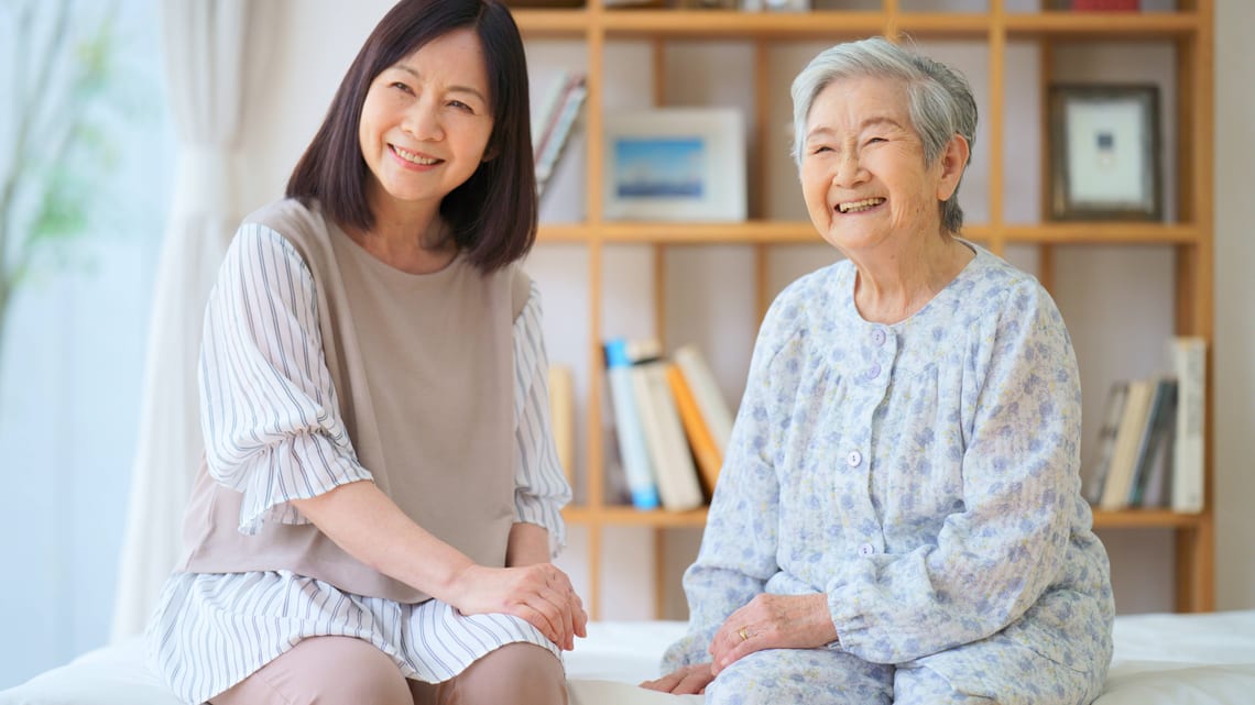 ベッドに並んで座り、笑顔で話す高齢のお母様と娘さんのイメージ