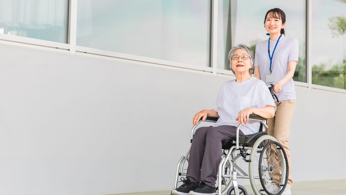病院の廊下で、車いすの高齢の女性と職員が散歩しているイメージ