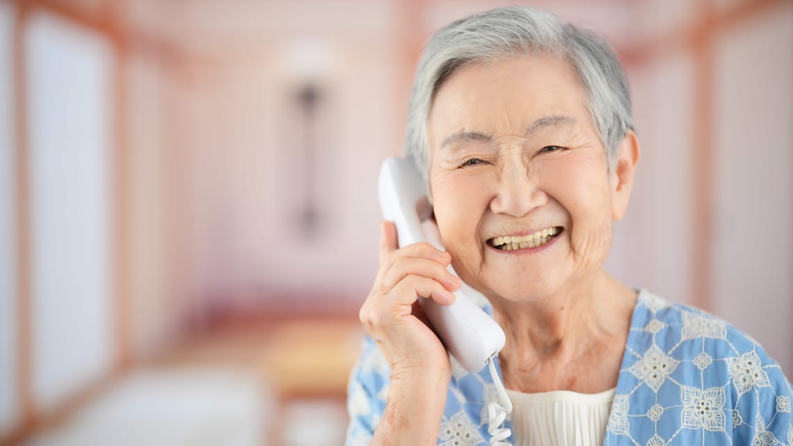 固定電話で、離れて暮らす家族と笑顔で話す高齢の女性のイメージ