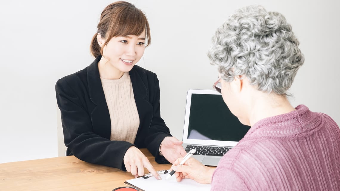 窓口で、職員の説明を聞きながら書類に記入する高齢の女性のイメージ