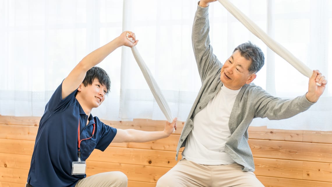 職員と一緒にタオル体操をする高齢の男性のイメージ