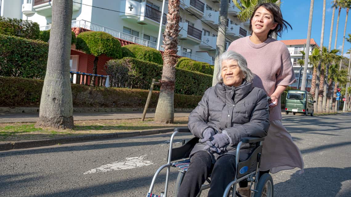 職員に車いすを押してもらい、外を散歩する高齢の女性のイメージ