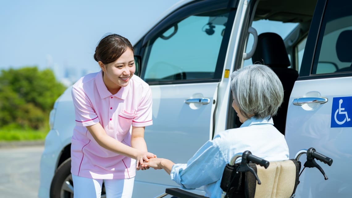 送迎の車の前で、車いすの方の手を取る介護職員のイメージ