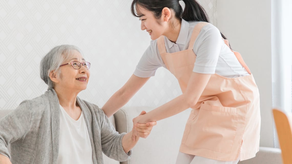 自宅で、ヘルパーに手を取られて立ち上がる高齢の女性のイメージ
