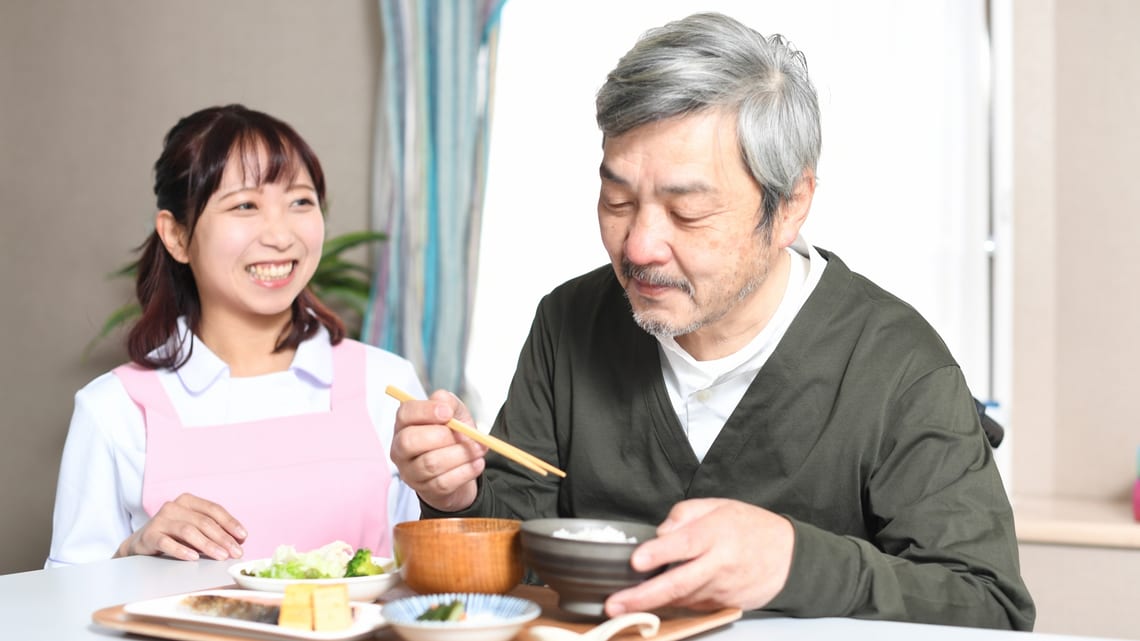 住まいの食堂で、職員と話しながら食事をとる高齢の男性のイメージ