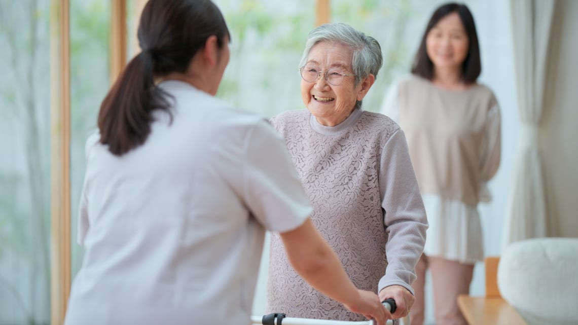 職員に付き添われて、歩行器で歩く練習をする高齢の女性と、見守る家族のイメージ