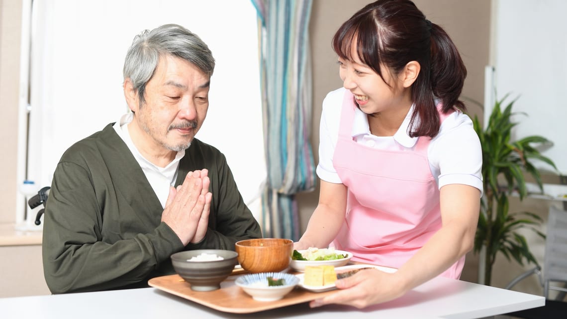 職員が笑顔で食事を運び、手を合わせる高齢の男性のイメージ