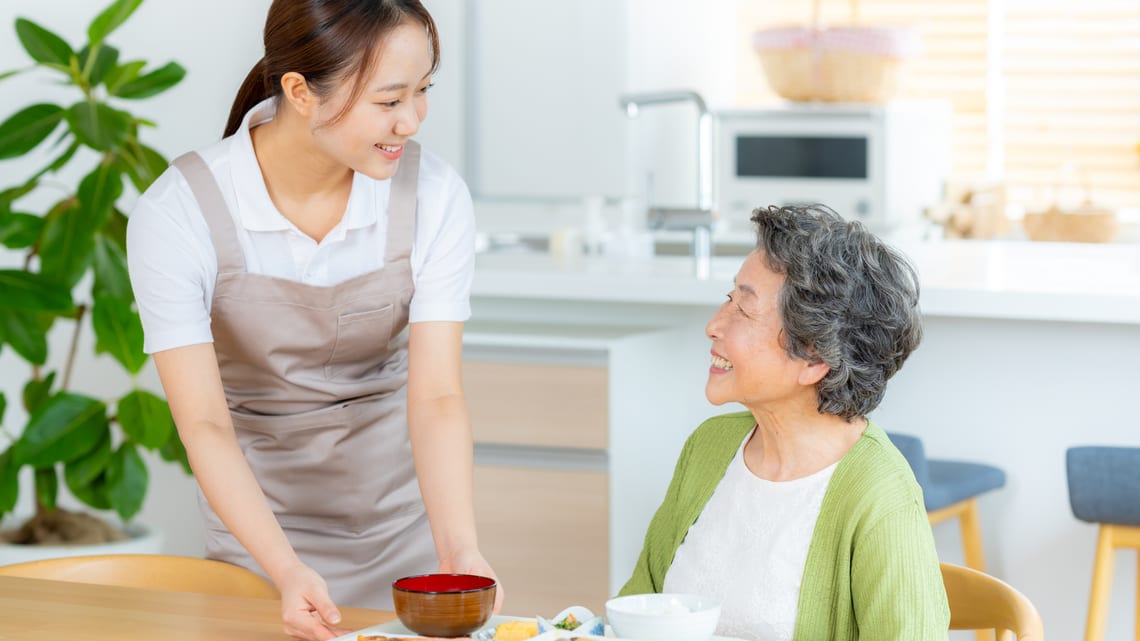 ご自宅で、ヘルパーが笑顔で食事を運ぶ高齢の女性のイメージ